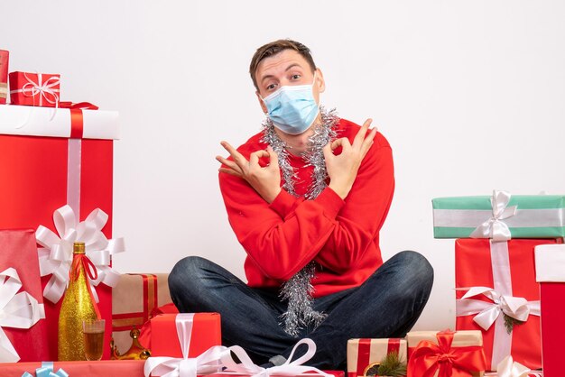 Vista frontal del joven en máscara sentado alrededor de regalos de Navidad en la pared blanca