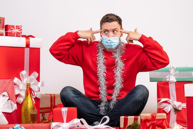 Vista frontal del joven en máscara sentado alrededor de regalos de Navidad en la pared blanca