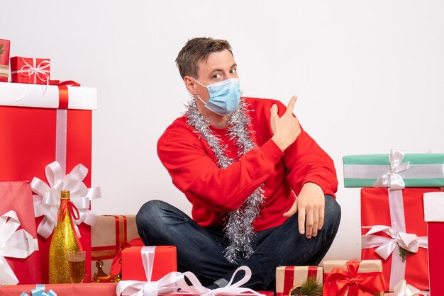 Vista frontal del joven en máscara sentado alrededor de regalos de Navidad en la pared blanca