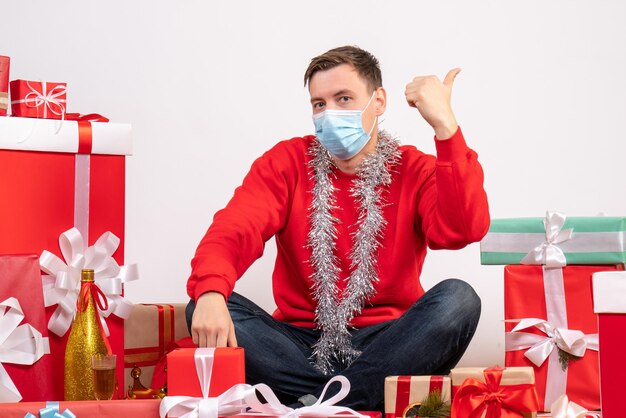 Vista frontal del joven en máscara sentado alrededor de regalos de Navidad en la pared blanca