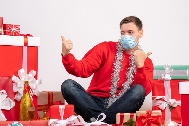 Vista frontal del joven en máscara sentado alrededor de regalos de Navidad en la pared blanca