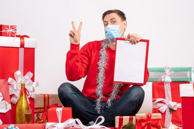 Vista frontal del joven en máscara sentado alrededor de regalos de Navidad en la pared blanca