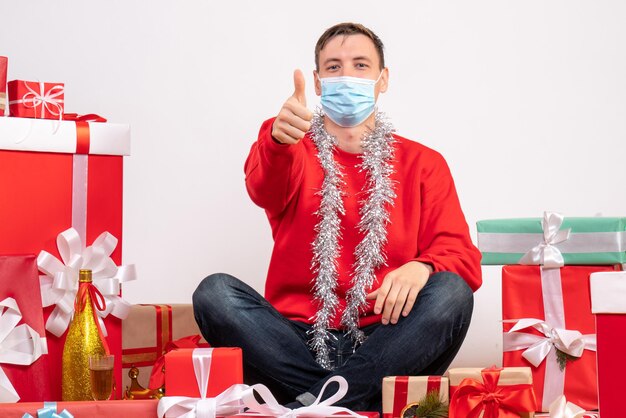 Vista frontal del joven en máscara sentado alrededor de regalos de Navidad en la pared blanca