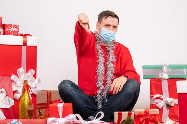 Vista frontal del joven en máscara sentado alrededor de regalos de Navidad en la pared blanca