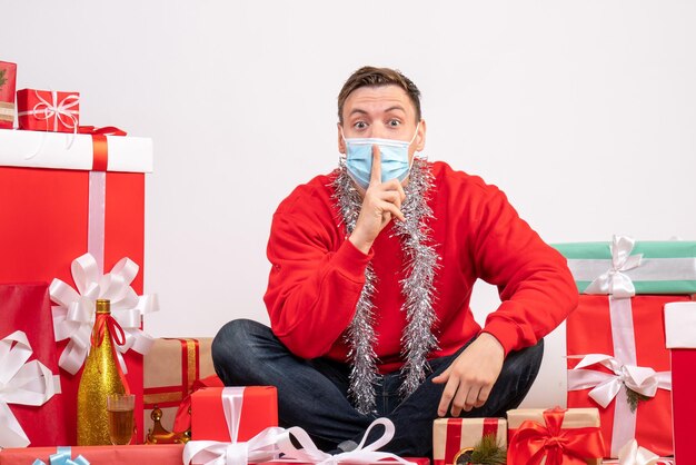 Vista frontal del joven en máscara sentado alrededor de regalos de Navidad en la pared blanca