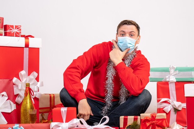 Vista frontal del joven en máscara sentado alrededor de regalos de Navidad en la pared blanca