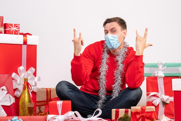 Vista frontal del joven en máscara sentado alrededor de regalos de Navidad en la pared blanca