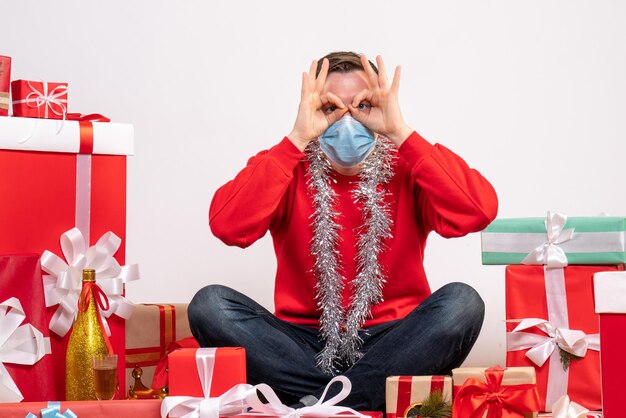 Vista frontal del joven en máscara sentado alrededor de regalos de Navidad en la pared blanca