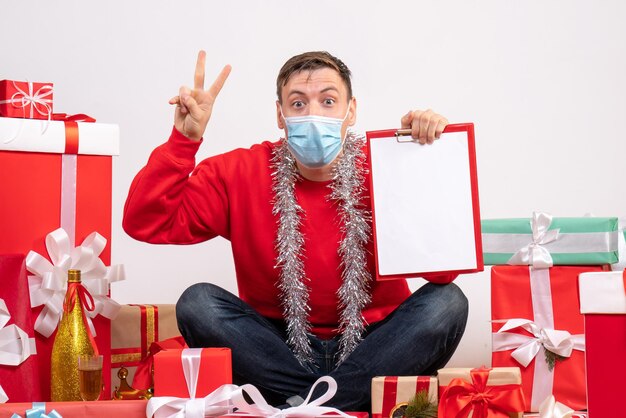 Vista frontal del joven en máscara sentado alrededor de regalos de Navidad en la pared blanca