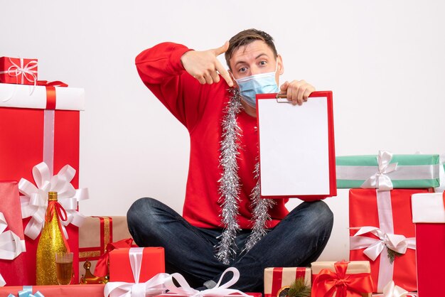 Vista frontal del joven en máscara sentado alrededor de regalos de Navidad en la pared blanca