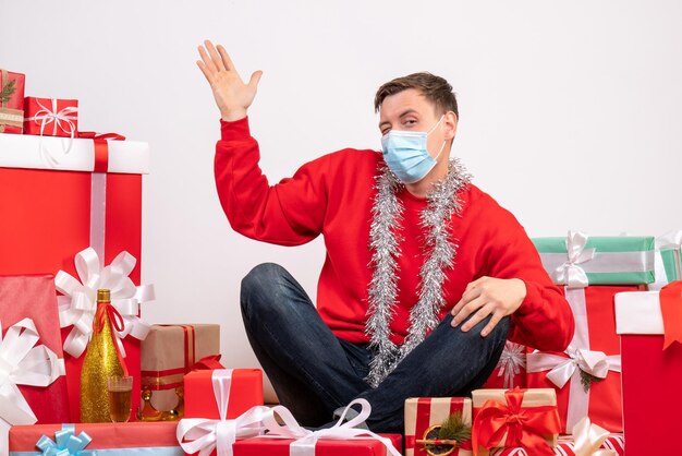 Vista frontal del joven en máscara sentado alrededor de regalos de Navidad en la pared blanca