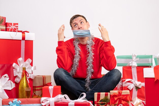 Vista frontal del joven en máscara sentado alrededor de regalos de Navidad en la pared blanca