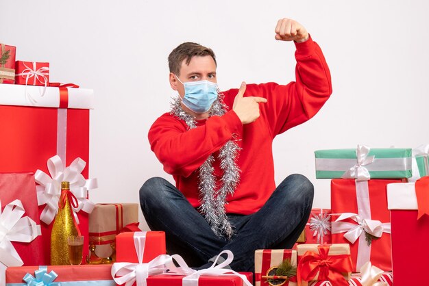 Vista frontal del joven en máscara sentado alrededor de regalos de Navidad en la pared blanca