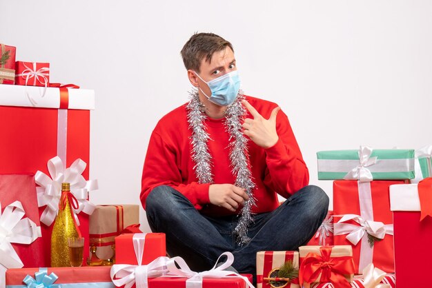 Vista frontal del joven en máscara sentado alrededor de regalos de Navidad en la pared blanca