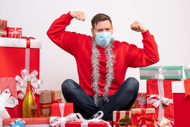 Vista frontal del joven en máscara sentado alrededor de regalos de Navidad en la pared blanca