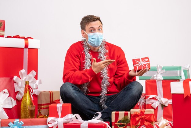 Vista frontal del joven en máscara sentado alrededor de regalos de Navidad en la pared blanca