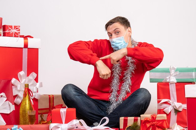 Vista frontal del joven en máscara sentado alrededor de regalos de Navidad en la pared blanca