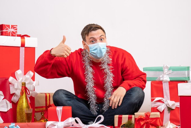 Vista frontal del joven en máscara sentado alrededor de regalos de Navidad en la pared blanca