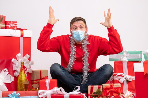 Vista frontal del joven en máscara sentado alrededor de regalos de Navidad en la pared blanca