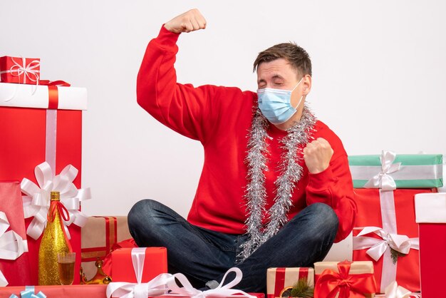 Vista frontal del joven en máscara sentado alrededor de regalos de Navidad en la pared blanca