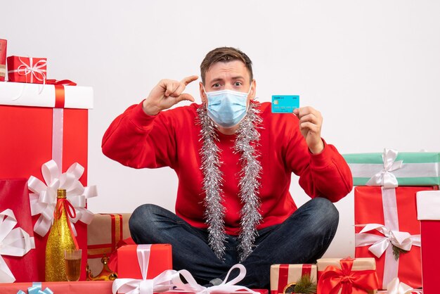 Vista frontal del joven con máscara sentado alrededor de regalos de Navidad con tarjeta bancaria en la pared blanca