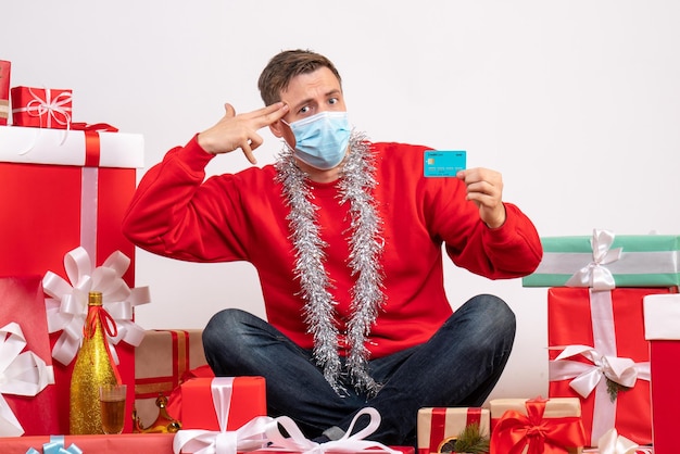 Vista frontal del joven con máscara sentado alrededor de regalos de Navidad con tarjeta bancaria en la pared blanca