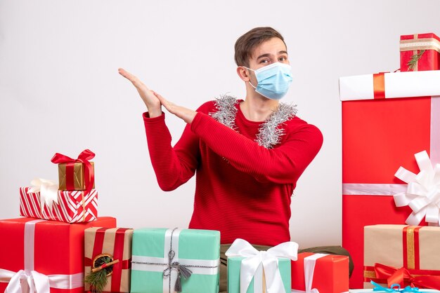 Vista frontal joven con máscara sentado alrededor de regalos de Navidad