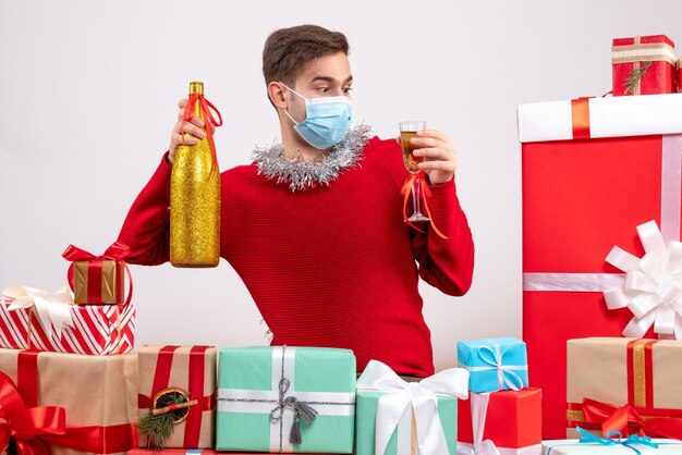 Vista frontal joven con máscara sosteniendo champán alrededor de regalos de Navidad