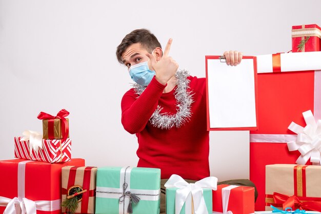 Vista frontal joven con máscara sosteniendo portapapeles sentado en el piso regalos de navidad