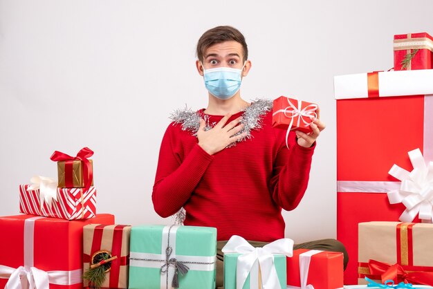 Vista frontal joven con máscara sosteniendo presente sentado alrededor de regalos de Navidad