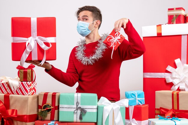 Vista frontal joven con máscara sosteniendo regalos de Navidad sentado alrededor de regalos de Navidad