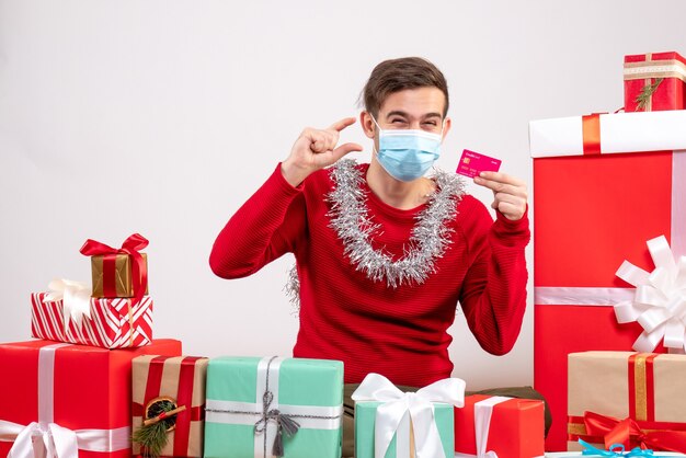 Vista frontal joven con máscara sosteniendo tarjeta sentado alrededor de regalos de Navidad en blanco