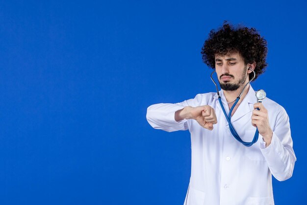 Vista frontal del joven médico masculino en traje médico con estetoscopio sobre fondo azul coronavirus salud covid-vacuna virus hospital droga pandemia