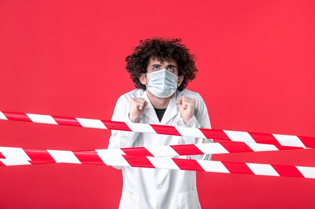 Vista frontal joven médico masculino en uniforme médico y máscara sobre fondo rojo aislamiento hospital covid- tira de advertencia de cuarentena de salud