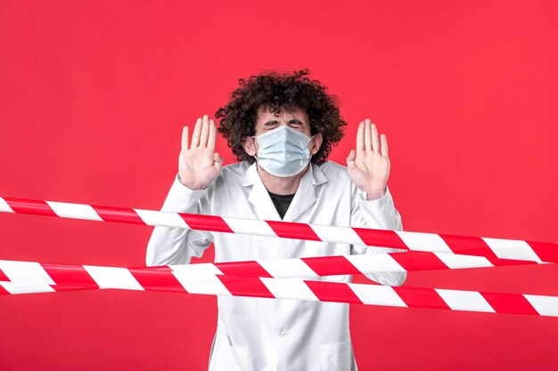 Vista frontal joven médico masculino en uniforme médico y máscara sobre fondo rojo aislamiento hospital droga covid- advertencia peligro de cuarentena