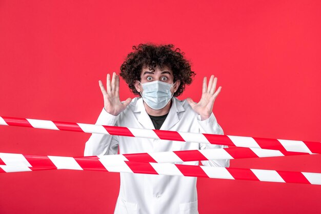 Vista frontal joven médico masculino en uniforme médico y máscara sobre fondo rojo aislamiento hospital droga covid peligro advertencia cuarentena