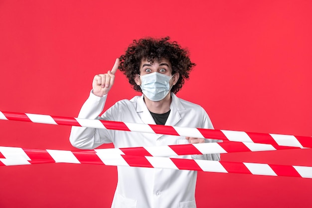 Vista frontal joven médico masculino en uniforme médico y máscara sobre fondo rojo peligro covid- hospital de tira de advertencia de cuarentena de salud