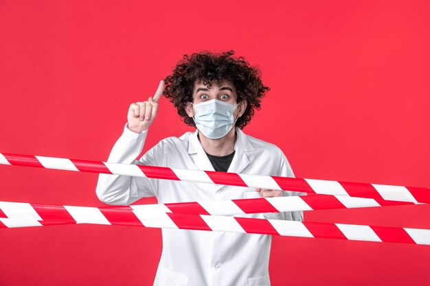 Vista frontal joven médico masculino en uniforme médico y máscara sobre fondo rojo peligro covid- hospital de tira de advertencia de cuarentena de salud