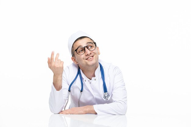 Vista frontal joven médico de pensamiento en traje médico sentado detrás del escritorio en la pared blanca