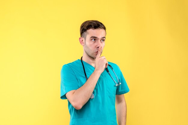 Vista frontal del joven médico pidiendo silencio en la pared amarilla