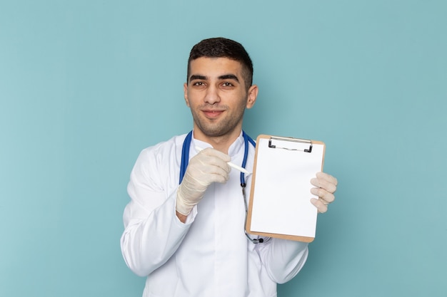 Vista frontal del joven médico en traje blanco con estetoscopio azul anotando notas