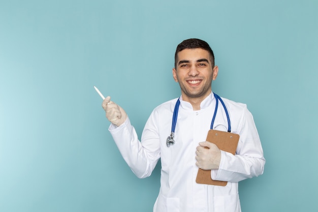 Vista frontal del joven médico en traje blanco con estetoscopio azul anotando notas