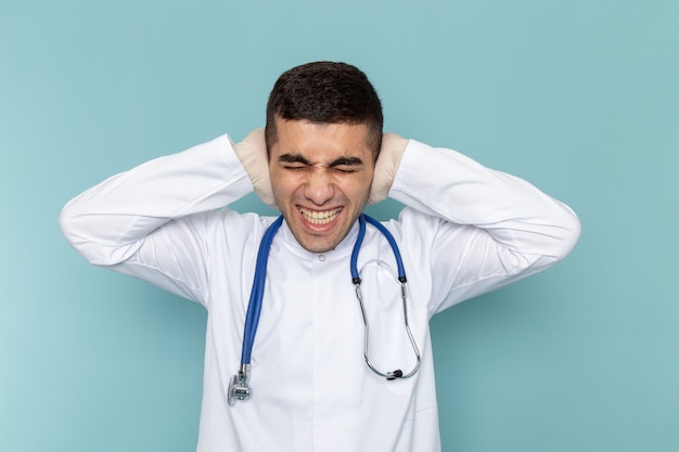 Vista frontal del joven médico en traje blanco con estetoscopio azul cubriendo sus oídos