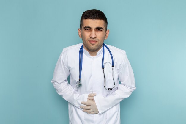 Vista frontal del joven médico en traje blanco con estetoscopio azul con dolor de estómago