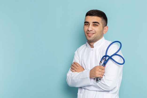 Vista frontal del joven médico en traje blanco con estetoscopio azul posando y sonriendo