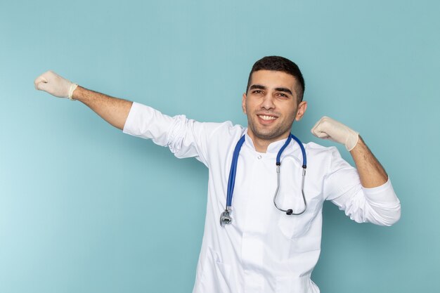 Vista frontal del joven médico en traje blanco con estetoscopio azul smilign y posando