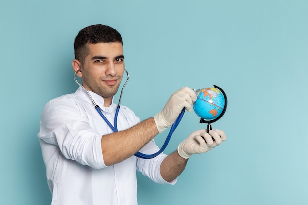 Vista frontal del joven médico en traje blanco con estetoscopio azul sonriendo y comprobando el globo