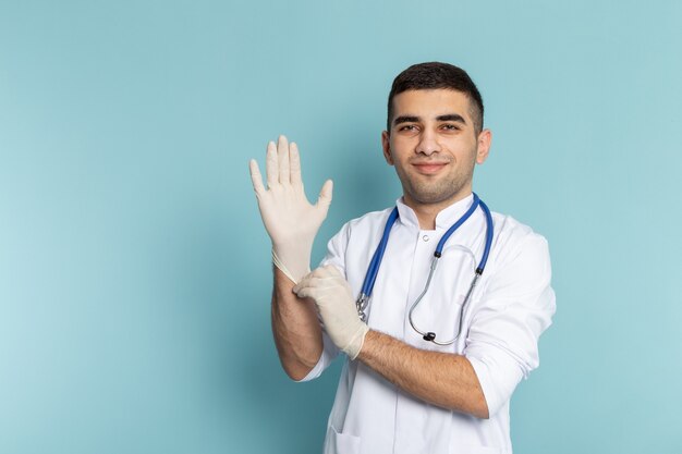 Vista frontal del joven médico en traje blanco con estetoscopio azul sonriendo con guantes