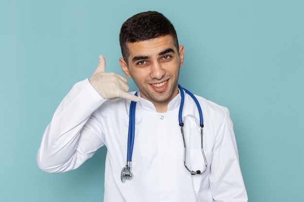 Vista frontal del joven médico en traje blanco con estetoscopio azul sonriendo y haciendo señal de teléfono