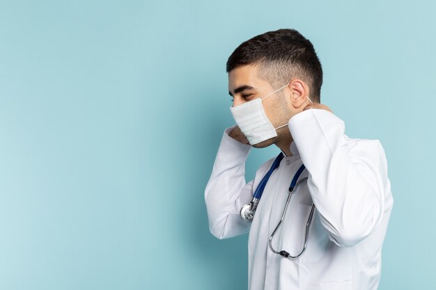 Vista frontal del joven médico en traje blanco con estetoscopio azul sonriendo con máscara